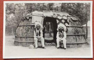 Item #H1164 RPPC, two Native Americans in full regalia sitting in front of a dwelling, Hayward WI...